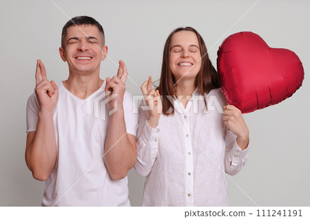 Hopeful couple standing with crossed fingers praying for good luck and fortune man and woman wearing white clothing holding heart shaped air balloon isolated over gray background Hopeful couple standing with crossed fingers praying for good luck and fortune man and woman wearing white clothing holding heart shaped air balloon isolated over gray background 111241191