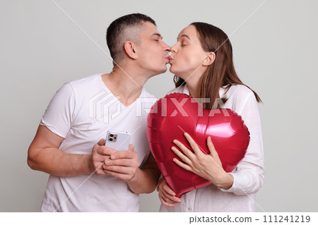 Romantic man and woman wearing white clothing holding heart shaped air balloon isolated over gray background using smartphone kissing celebrating anniversary or Valentines Day 111241219