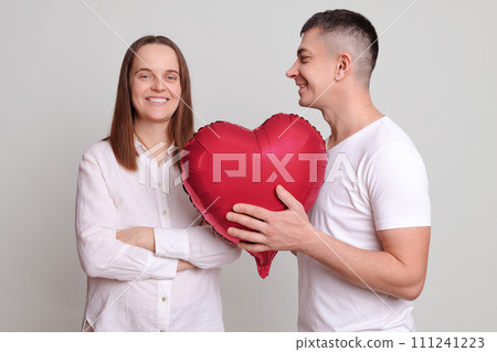 Smiling couple celebrating Valentines Day man giving present to smiling cheerful woman with heart shaped balloon expressing romantic feelings isolated over grey background Smiling couple celebrating Valentines Day man giving present to smiling cheerful woman with heart shaped balloon expressing romantic feelings isolated over grey background 111241223