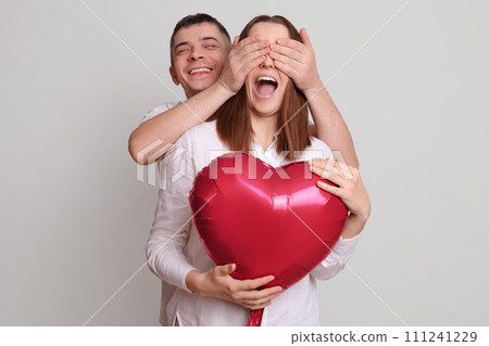 couple man and woman with heart shaped balloon isolated over grey background husband covering wife eyes with hands having fun together enjoying happy moments of Valentines Day celebration 111241229