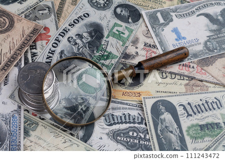 Old west period US banknotes and silver certificates with silver dollar coins and magnifying glass 111243472