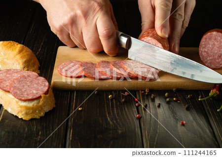 The cook cuts salami sausage on a wooden cutting board. The concept of preparing sandwiches for a snack in a restaurant kitchen 111244165