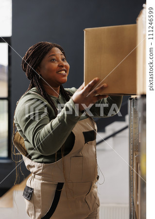 Stockroom worker taking out cardboard box from shelves, preparing customers online orders in warehouse. African american supervisor wearing industrial overall working at products delivery in Stockroom worker taking out cardboard box from shelves, preparing customers online orders in warehouse. African american supervisor wearing industrial overall working at products delivery in 111246599
