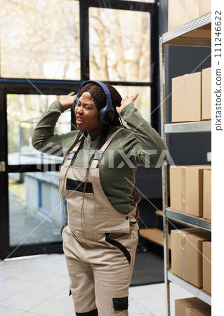 Small business employee dancing during work break in warehouse, listening music and having fun before start working at storage room inventory. African american worker wearing headphones Small business employee dancing during work break in warehouse, listening music and having fun before start working at storage room inventory. African american worker wearing headphones 111246622