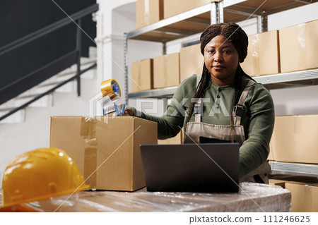 African american employee wearing industrial overall working in storehouse, preparing customers orders for delivery. Storage room employee checking shipping detalis on laptop computer, portrait African american employee wearing industrial overall working in storehouse, preparing customers orders for delivery. Storage room employee checking shipping detalis on laptop computer, portrait 111246625