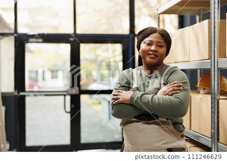 Smiling stockroom manager standing with arm crossed, posing with confidence in warehouse. African american employee wearing industrial overall while working at customers online orders 111246629