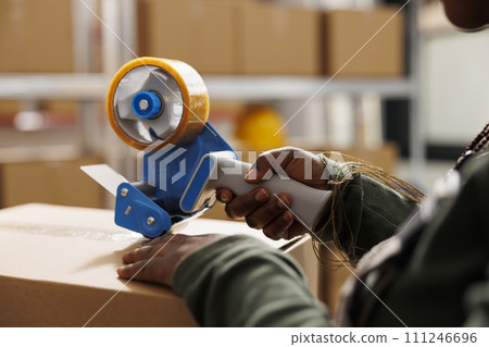 Storehouse worker using adhesive tape to pack clients orders, preparing packages for delivery in storehouse. African american employee wearing industrial overall during merchandise inventory. Close up Storehouse worker using adhesive tape to pack clients orders, preparing packages for delivery in storehouse. African american employee wearing industrial overall during merchandise inventory. Close up 111246696