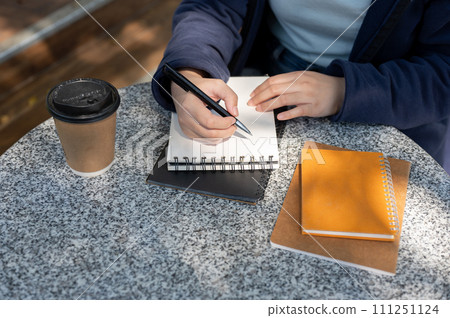 Woman taking notes in a spiral notepad while sitting at an outdoor table, working remotely at a cafe Woman taking notes in a spiral notepad while sitting at an outdoor table, working remotely at a cafe 111251124
