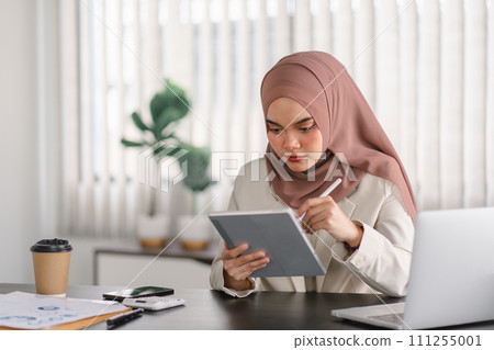 Asian Muslim businesswoman using a digital tablet while working indoors. 111255001