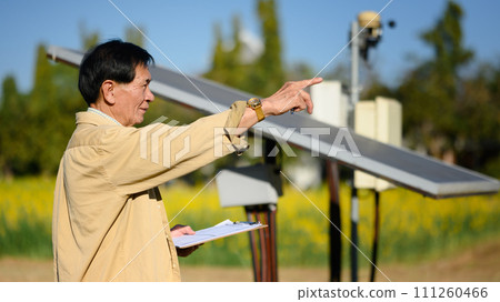 Smiling senior man holding clipboard standing beside solar panel at the cornfield. Renewable energy and agriculture concept 111260466