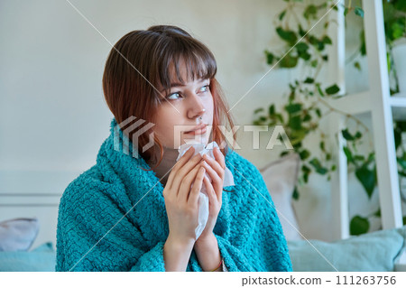 Young sick woman sitting under warm blanket sneezing coughing in handkerchief Young sick woman sitting under warm blanket sneezing coughing in handkerchief 111263756