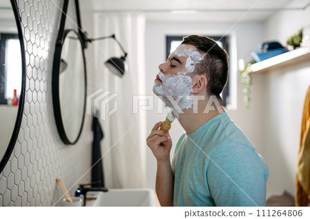 Young man with down syndrome learning how to shave, applying shaving foam all over his face, looking at mirror. Young man with down syndrome learning how to shave, applying shaving foam all over his face, looking at mirror. 111264806