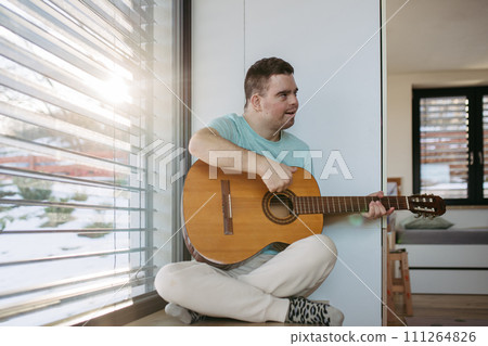 Young man with down syndrome playing acoustic guitar, sitting by window, holding and strumming guitar, making music. Young man with down syndrome playing acoustic guitar, sitting by window, holding and strumming guitar, making music. 111264826