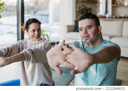 Young man with Down syndrome exercising at home with his mother on fitness ball. Workout routine for disabled man. Young man with Down syndrome exercising at home with his mother on fitness ball. Workout routine for disabled man. 111264831