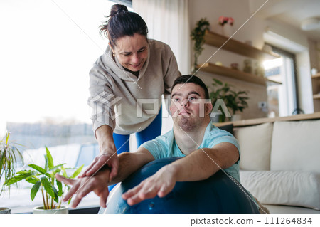 Young man with Down syndrome exercising at home with his mother on fitness ball. Workout routine for disabled man. Young man with Down syndrome exercising at home with his mother on fitness ball. Workout routine for disabled man. 111264834