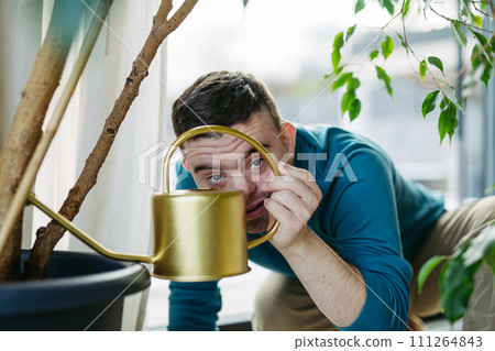 Young man with Down syndrome taking care of indoor plants, watering them, looking at camera through handle of a metal watering can. 111264843