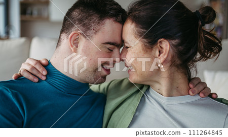 Portrait of young man with Down syndrome with his mother at home, holding, touching with foreheads. Concept of love and parenting disabled child. Banner with copyspace. Portrait of young man with Down syndrome with his mother at home, holding, touching with foreheads. Concept of love and parenting disabled child. Banner with copyspace. 111264845