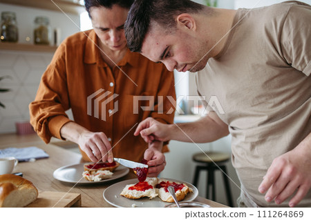 Young man with Down syndrome preparing breakfast with his mother at home. Morning routine for man with Down syndrome. Young man with Down syndrome preparing breakfast with his mother at home. Morning routine for man with Down syndrome. 111264869