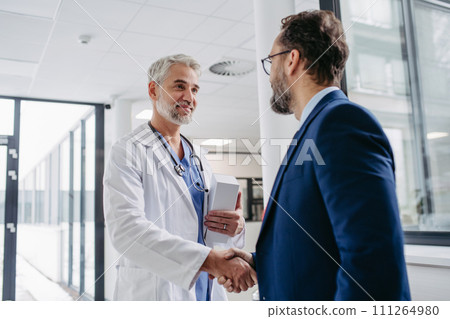Pharmaceutical sales representative presenting new medication to doctor in medical building, shaking hands. 111264980