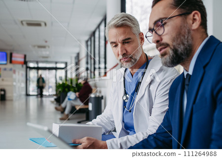 Pharmaceutical sales representative talking with doctor in medical building, presenting new medication on tablet. Hospital director, manager in modern clinic with surgeon. 111264988