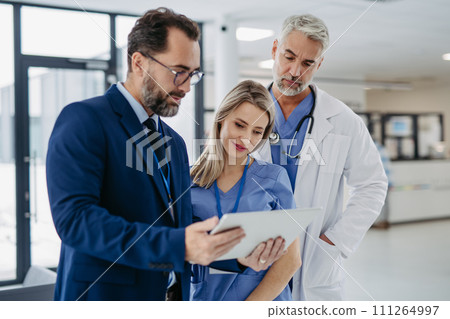 Pharmaceutical sales representative talking with doctors in medical building, presenting new medication on tablet. Hospital director, manager in modern clinic with doctor and nurse. 111264997