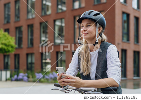 Beautiful middle-aged woman commuting through the city by bike, holding smartphone. Female city commuter traveling from work by bike after long workday. 111265185