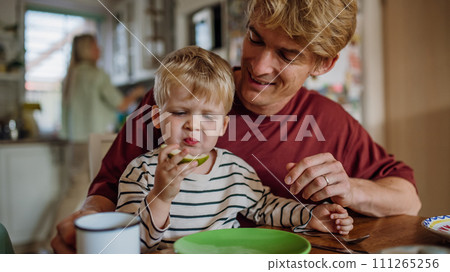 Father helping toddler to eat breakfast, home kitchen. Healthy breakfast or snack before kindergarden and work. Father helping toddler to eat breakfast, home kitchen. Healthy breakfast or snack before kindergarden and work. 111265256
