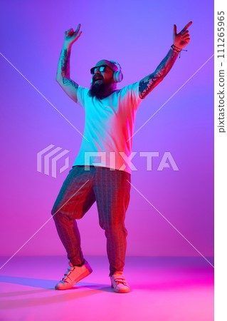 Full-length image of bearded bald man in casual clothes, with tattoos, listening to music in headphones against gradient purple background in neon light 111265965