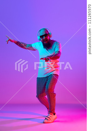 Full-length image of bearded bald man in casual clothes, with tattoos, listening to music in headphones against gradient purple background in neon light Full-length image of bearded bald man in casual clothes, with tattoos, listening to music in headphones against gradient purple background in neon light 111265970