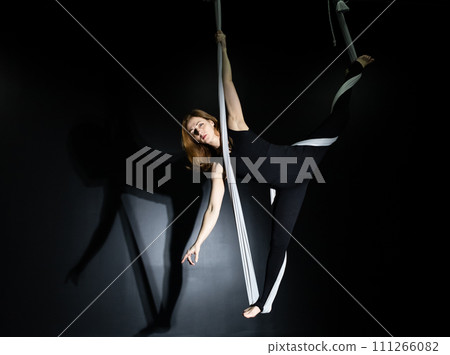 Fly yoga. Girl in a white hammock on a black background shows aerial acrobatics. Gymnastics, circus, under dome. Play of light and shadow creates feeling of a theatrical performance. Sports activity 111266082