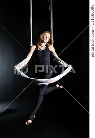 Fly yoga. Girl in a white hammock on a black background shows aerial acrobatics. Gymnastics, circus, under dome. Play of light and shadow creates feeling of a theatrical performance. Sports activity 111266098