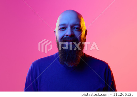 Gel portrait of bearded bald man in his 30s wearing blue sweater, smiling, looking in camera against pink background in neon light 111266123