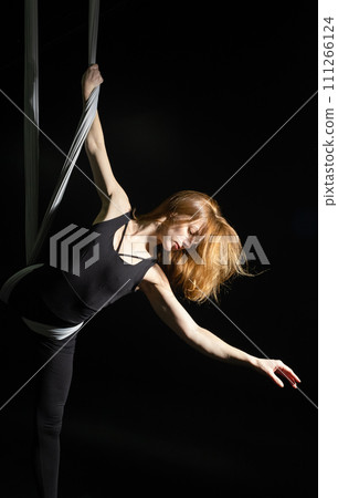 Fly yoga. Girl in a white hammock on a black background shows aerial acrobatics. Gymnastics, circus, under dome. Play of light and shadow creates feeling of a theatrical performance. Sports activity 111266124