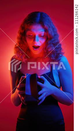 Present surprise. Special gift. Shocked delighted young woman with glowing face opening paper package isolated on pink spotlight background. Present surprise. Special gift. Shocked delighted young woman with glowing face opening paper package isolated on pink spotlight background. 111266142