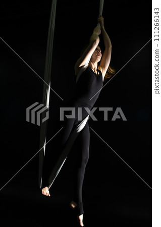 Fly yoga. Girl in a white hammock on a black background shows aerial acrobatics. Gymnastics, circus, under dome. Play of light and shadow creates feeling of a theatrical performance. Sports activity Fly yoga. Girl in a white hammock on a black background shows aerial acrobatics. Gymnastics, circus, under dome. Play of light and shadow creates feeling of a theatrical performance. Sports activity 111266143