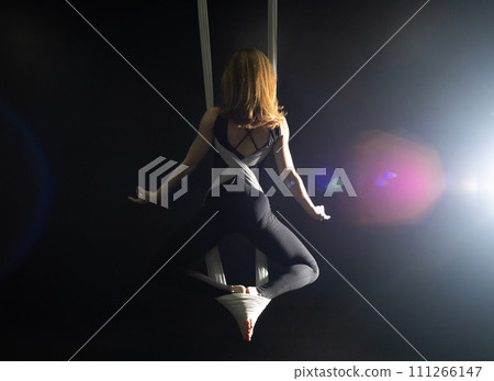 Fly yoga. Girl in a white hammock on a black background shows aerial acrobatics. Gymnastics, circus, under dome. Play of light and shadow creates feeling of a theatrical performance. Sports activity Fly yoga. Girl in a white hammock on a black background shows aerial acrobatics. Gymnastics, circus, under dome. Play of light and shadow creates feeling of a theatrical performance. Sports activity 111266147