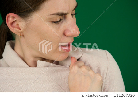the woman is cold, she has pulled a beige jacket around her, she is upset, sadly dissatisfied, on a green background, chroma key in her ears, earrings, big nose, mouth slightly open, eyes narrowed 111267320