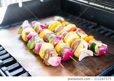 Skewered beef and fresh veggies sizzle on a copper grill mat over a gas outdoor grill. 111267575