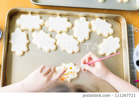 A heartwarming scene of a little girl carefully writing 'Sorry' on sugar cookies with food coloring, the cookies beautifully flooded with white royal icing. 111267721