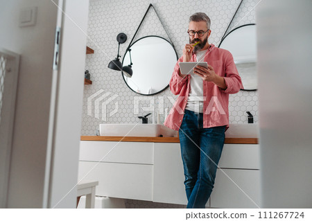 Man shopping online while brushing his teeth in bathroom. Holding tablet, scrolling and buying in eshop, toothbrush in mouth. Man shopping online while brushing his teeth in bathroom. Holding tablet, scrolling and buying in eshop, toothbrush in mouth. 111267724