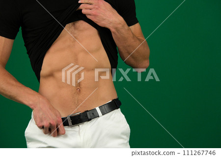 a man has beautiful abs, pumped up six-pack, white jeans, a black belt, a black T-shirt on a green background, unrecognizable people lift up their clothes, show their belly, sport, body beauty a man has beautiful abs, pumped up six-pack, white jeans, a black belt, a black T-shirt on a green background, unrecognizable people lift up their clothes, show their belly, sport, body beauty 111267746