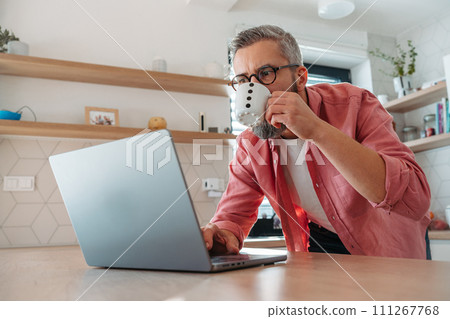 Mature man working from home on laptop, standing by kitchen island, with cup of coffee, holding smartphone. Concept of remote work from home. Hygge at work. Mature man working from home on laptop, standing by kitchen island, with cup of coffee, holding smartphone. Concept of remote work from home. Hygge at work. 111267768