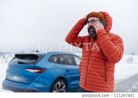 Angry man standing by electric car, battery run out of power before reaching destination. Man phone calling for help waiting for breakdown service car, tow truck. 111267881