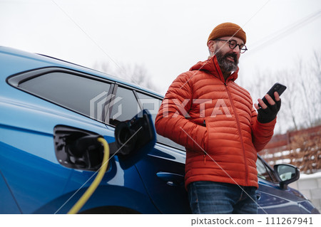 Man charging electric car during cold snowy day, using electric vehicle charging app, checking battery life, energy consumption on smart phone. Charging and driving electric vehicles during winter Man charging electric car during cold snowy day, using electric vehicle charging app, checking battery life, energy consumption on smart phone. Charging and driving electric vehicles during winter 111267941