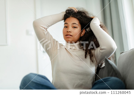 Portrait of beautiful curvy woman, sitting on bed, holding her hair, looking at camera. 111268045