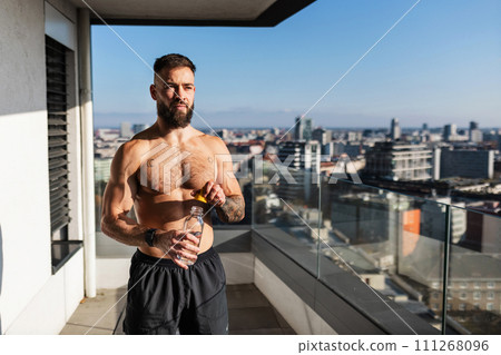 Handsome man with bare chest standing on balcony and enjoying beautiful view on city. Concept of selfcare and enjoying life for young people. Handsome man with bare chest standing on balcony and enjoying beautiful view on city. Concept of selfcare and enjoying life for young people. 111268096