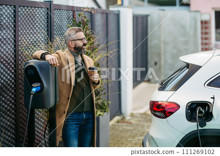 Portrait of businessman waiting while electric car is charging, leaning against vehicle, scrolling on his smartphone, and drinking coffee. An electric vehicle charging station. 111269102