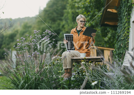 Woman in garden holding solar panel and tablet. Looking for solar panels grants, funds for homeowners. Solar energy and sustainable lifestyle for family in house. 111269194