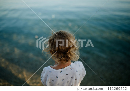 Rear view of small girl with blonde hair standing at beach looking at sea. Rear view of small girl with blonde hair standing at beach looking at sea. 111269222
