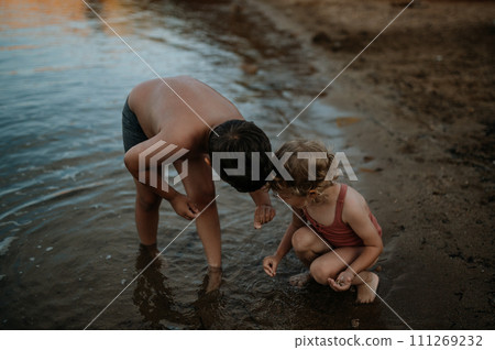 Brother helping sister seraching for shells in sand. Small girl in swimsuit playing at beach, crouching in water. 111269232